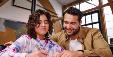 A father sits with his daughter at her desk, helping her with her work